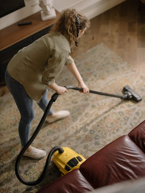 A professional cleaner dressed in a white protective suit, face mask, gloves, and shoe covers is performing surface cleaning on a beige carpet in a well-lit, modern living room. The cleaner is using a